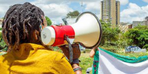 Call to Action AI genrated by Mwendwa Kivuva using DALL·E 2023-03-01 13.45.55 - A woman holding a loud speaker in Nairobi city calling a group of civil society activists to a town hall meeting